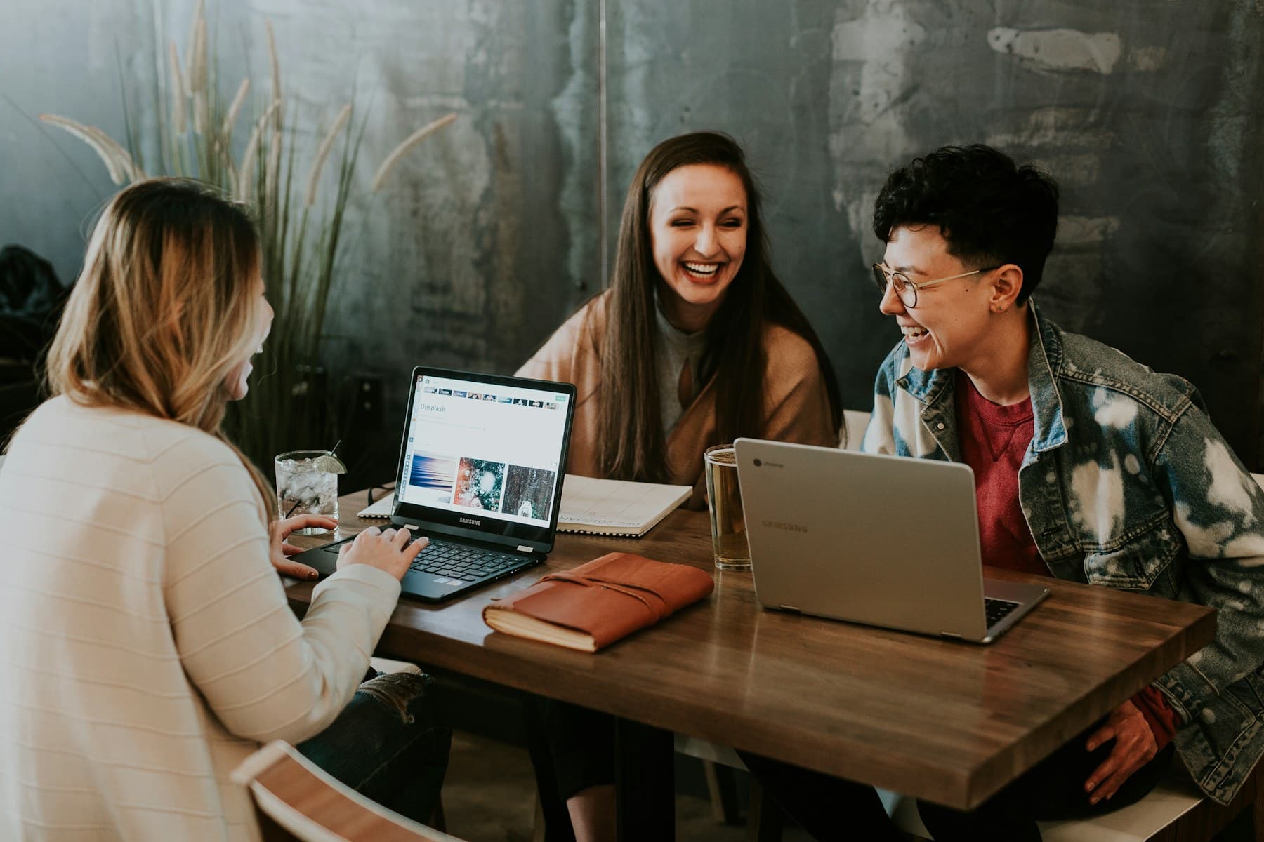 Three teammates laughing together over laptops at a wooden table